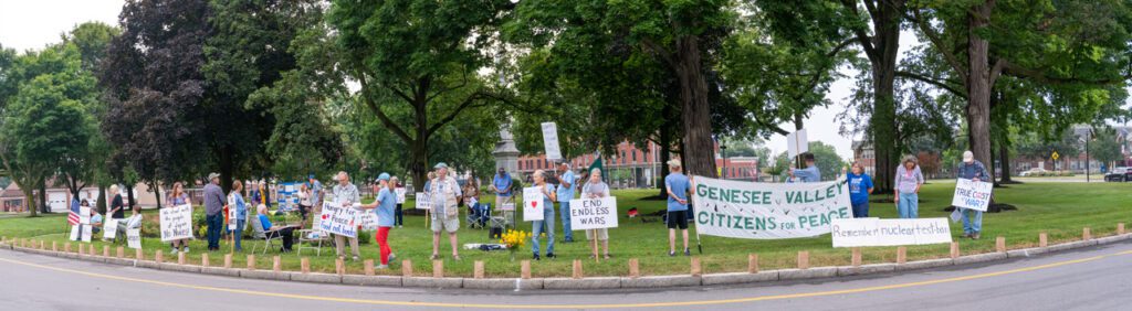 A group of people stand in a line holding various protest signs about peace and anti-war sentiments, in a park setting with trees and grass. A prominent banner reads "Genesee Valley Citizens for Peace."