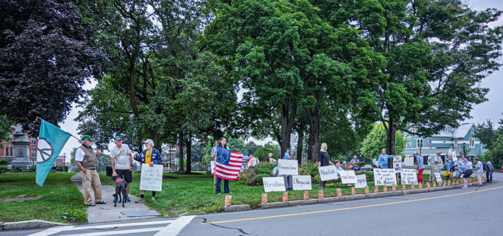 A group of people are gathered in a park holding signs advocating for the abolition of nuclear weapons. One person holds an American flag, while another holds a banner with a peace symbol. The scene is set against a background of green trees and a sidewalk, with some participants standing and others seated.