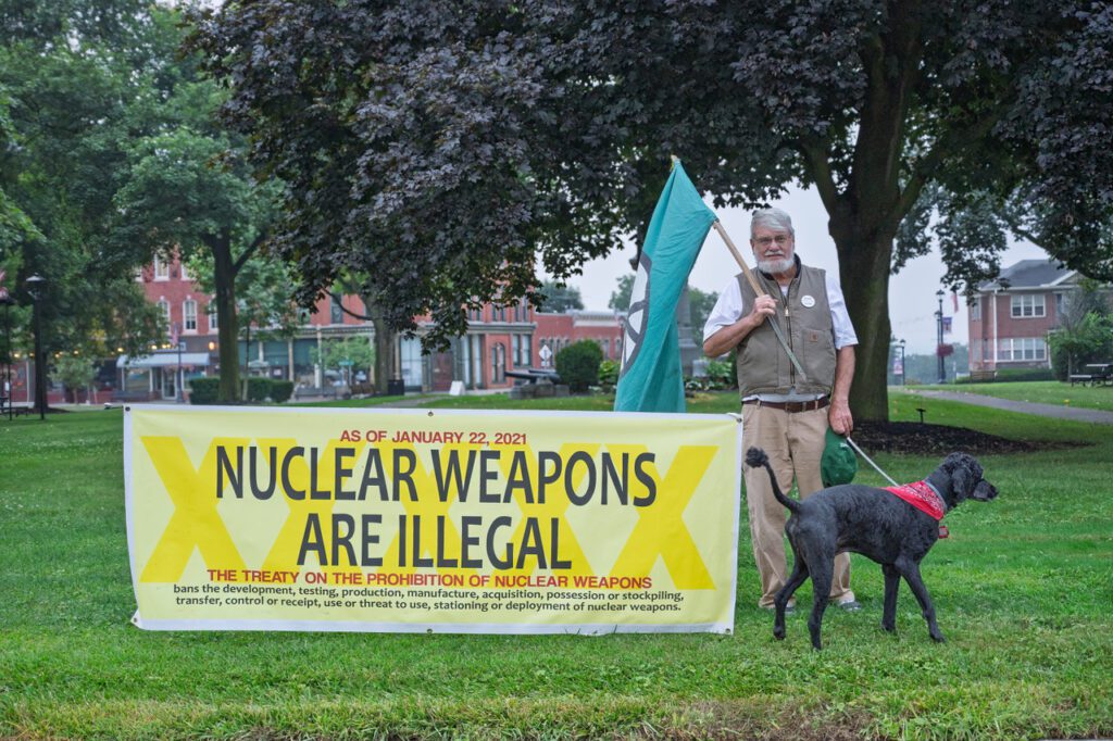 A man holding a green flag stands next to a yellow banner that reads 'Nuclear Weapons Are Illegal' in a park, alongside a dog wearing a red bandana.