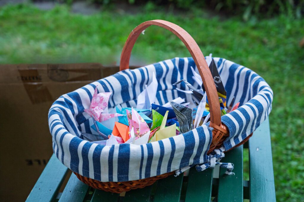 A wicker basket lined with blue and white striped fabric, containing colorful origami figures, placed on a green wooden surface with grass in the background.