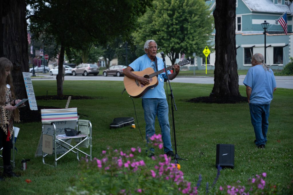 A man playing an acoustic guitar and singing into a microphone in a park setting. There is a folding chair with a striped bag beside him, and another person standing nearby. The background features trees, grass, and parked cars on a street.