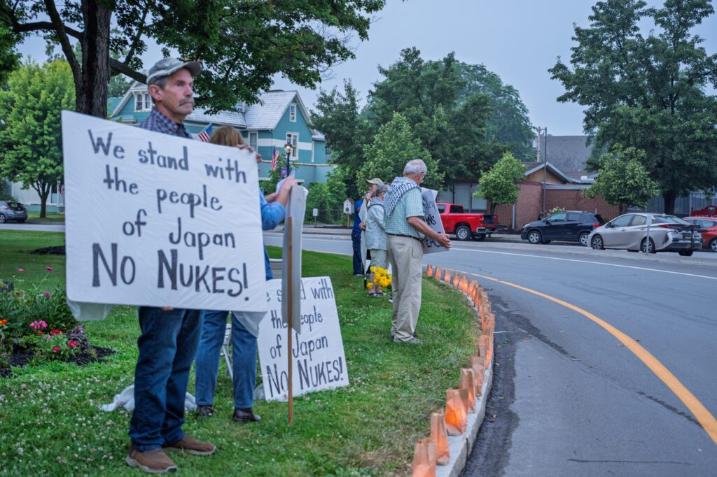 A group of people standing along a roadside holding signs that read "We stand with the people of Japan No Nukes." They are next to a row of paper lanterns lining the curb, with trees and a few parked cars in the background.