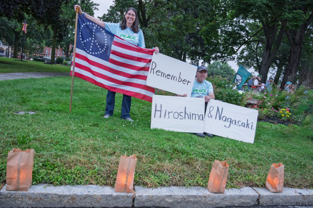 Two people hold a peace flag and signs reading "Remember Hiroshima & Nagasaki" at a candlelit vigil in a park.