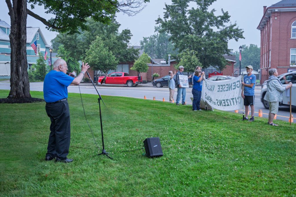 Dr. Matlin speaks into a microphone on a grassy area while a small group of people stand nearby holding protest signs. One sign reads 'What's the true cost of war?' and another banner says 'Sensible values, citizens for peace.' In the background, there are parked cars, trees, and residential buildings.